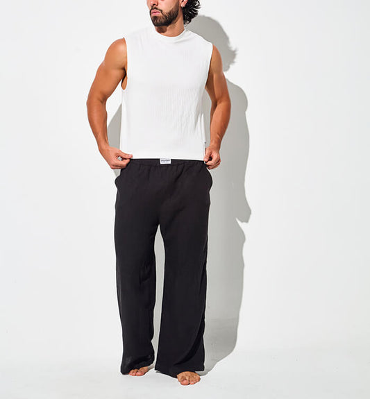 Man wearing a sleeveless white top and loose black pants, standing barefoot.